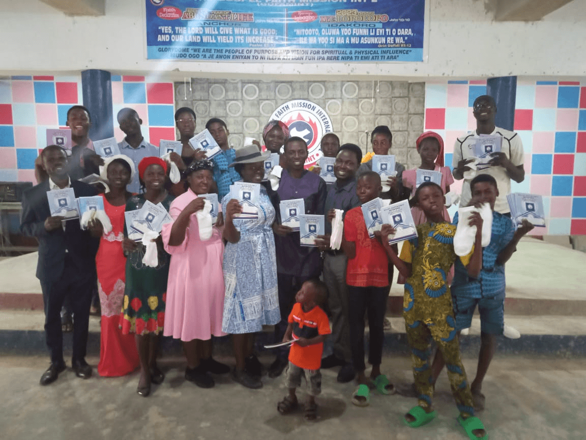 A large group of children, teenagers and adults standing together, each holding exercise books and a pair of white socks, under a church banner.