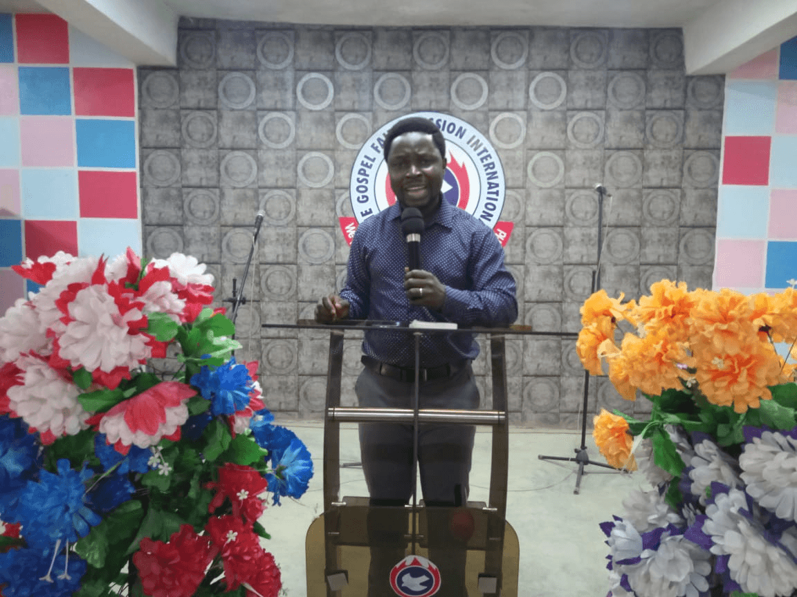 A man speaking into a microphone from a lectern, flanked by flower arrangements.
