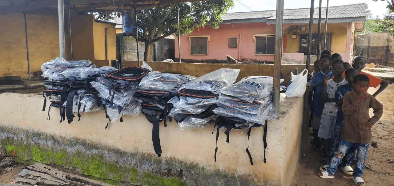 Dozens of new school bags in packaging, lined up along a low wall, with a few children waiting beside them.